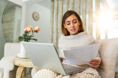 Indoor shot of casually dressed young woman holding papers in her hands, calculating and paying bills at home. Happy beautiful woman sitting on the sofa with laptop calculating home finances