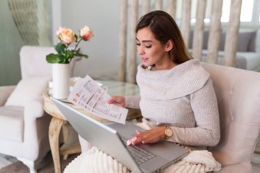 Indoor shot of casually dressed young woman holding papers in her hands, calculating and paying bills at home. Happy beautiful woman sitting on the sofa with laptop calculating home finances