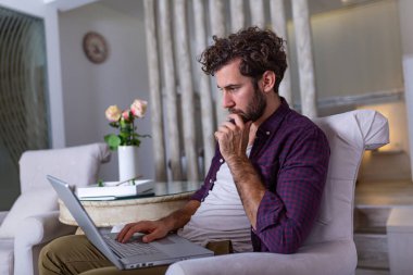 Ciddi yüzlü, ciddi işportacı evde oturmuş, dizüstü bilgisayarını projesi için kullanıyor. Ev konseptinden bağımsız olarak çalışan iş adamı cep telefonu ve dizüstü bilgisayarında istatistikleri kontrol ediyor