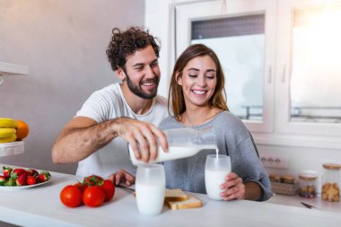 Genç ve neşeli bir çift mutfakta kahvaltı yaparken eğleniyor. Neşeli genç adam, güzel karısına sarılıyor, kahvaltıda sütünü döküyor.