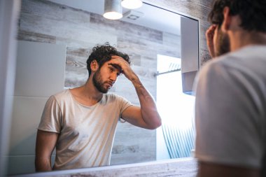 Yakışıklı genç adam elleriyle saçına dokunuyor ve evdeki banyoda tımar ediyor. Beyaz metroseksüel adam saç dökülmesinden endişeleniyor ve aynaya bakıyor..