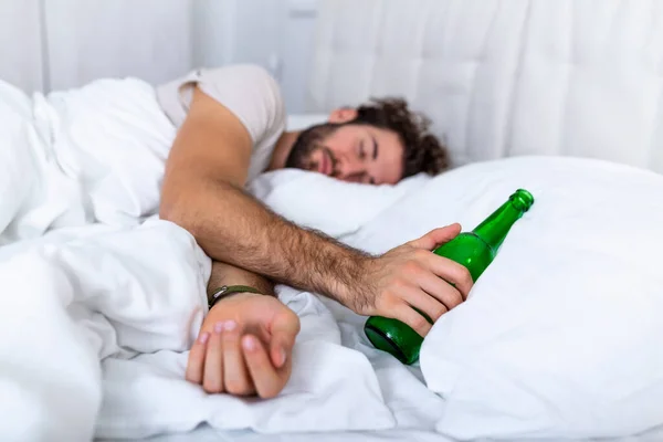Drunk man in the bed and sad place and an alcohol bottle in his hand. Young man lying in bed deadly drunken holding near-empty bottle of booze.