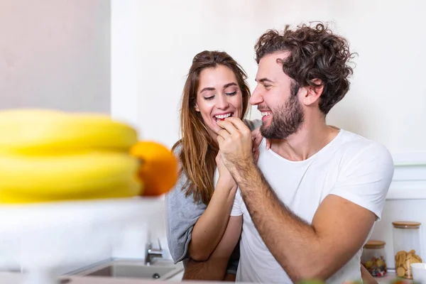 Beautiful young couple is feeding each other and smiling while cooking in kitchen at home. Happy sporty couple is preparing healthy food on light kitchen. Healthy food concept.