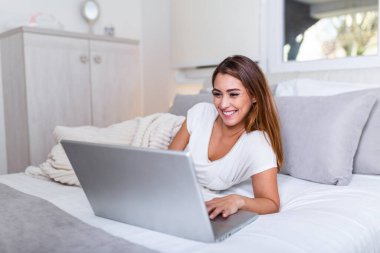 Happy casual beautiful woman working on a laptop laying on the bed in the house. Freelancer woman working on her laptop at home