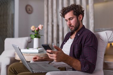 Genç yakışıklı bir adam, modern evindeki koltukta otururken arkadaşlarıyla video görüşmesi yapıyor. Mutlu iş adamları kavramı. İş adamı cep telefonu ve laptop istatistiklerini kontrol ediyor.