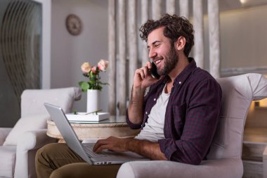 Genç, çekici, gülümseyen adam dizüstü bilgisayarına bakıyor ve konuşuyor. Cep telefonu, rahat koltukta oturuyor, günlük kıyafetler giyiyor. Ev konseptinden bağımsız iş