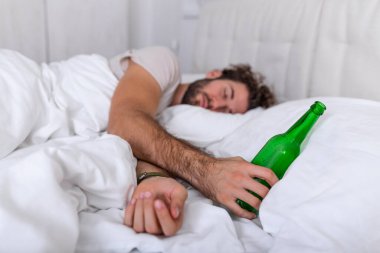 Yatakta sarhoş bir adam, üzgün bir yer ve elinde bir alkol şişesi. Yatakta yatan genç adam sarhoş halde elinde boş bir şişe içki tutuyordu..