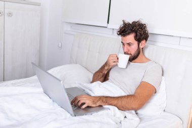 Kafkasyalı genç bir adamın sabah kahvesini içtiği, yatağında oturduğu, laptopuyla çalıştığı ve kahve içtiği, incelenmeye hazırlandığı, internette sörf yapmak için dizüstü bilgisayarını kullandığı görüntüsü..
