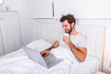 Kafkasyalı genç bir adamın sabah kahvesini içtiği, yatağında oturduğu, laptopuyla çalıştığı ve kahve içtiği, incelenmeye hazırlandığı, internette sörf yapmak için dizüstü bilgisayarını kullandığı görüntüsü..