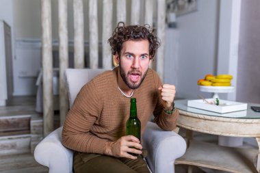 Evdeki kanepede spor maçı izlerken tezahürat yapan heyecanlı genç adam. Çekici, mutlu adam futbol seyrediyor, en sevdiği takımın zaferini kutluyor, bağırıyor, bira içiyor. .