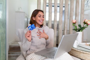 Genç ve güzel bir kadın internetten alışveriş malzemeleri bulduğu için mutlu. Güzel kız evde online alışveriş için dizüstü bilgisayar kullanıyor.