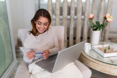 Kredi kartı tutan ve dizüstü bilgisayar kullanan genç bir kadın. İnternetten alışveriş konsepti. Mutlu kadın evde online alışveriş yapıyor.