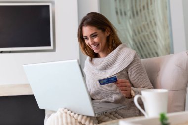 Genç ve güzel bir kadın internetten alışveriş malzemeleri bulduğu için mutlu. Güzel kız evde online alışveriş için dizüstü bilgisayar kullanıyor.