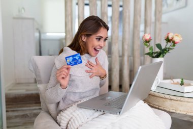 Genç ve güzel bir kadın internetten alışveriş malzemeleri bulduğu için mutlu. Güzel kız evde online alışveriş için dizüstü bilgisayar kullanıyor.