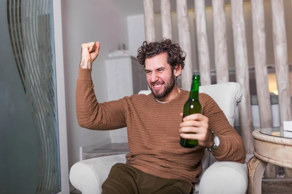 テレビでサッカーの試合を見ている熱狂的なスポーツファンの男を祝う 彼の好きなチームの勝利を祝いビールを飲みリビングルームに座って — ストック写真