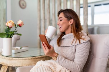 Mutlu gülümseyen güzel kız rahat evinde akıllı telefon kullanıyor, çekici genç kadın sosyal ağda arkadaşlarıyla sohbet ediyor ve evde kanepede oturup kahve içiyor.