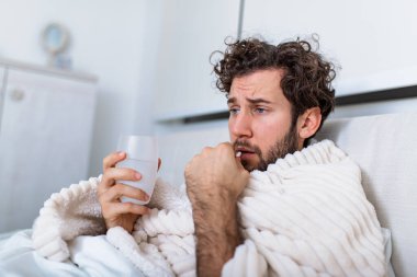 Hasta, ziyan olmuş, pijama giymiş, soğuk algınlığı virüsü kapmış hasta bir adam. Sağlık sigortası konseptinde ilaç tabletleri var. Bir bardak suyla hap içiyor.