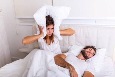 Man snoring while his wife is covering ears with pillow. Woman suffers from her male partner snoring in bed. Noise concept. Real people. Young irritated woman lying in bed with snoring husband clipart