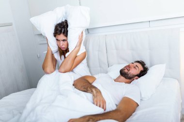 Young couple have problem with man's snoring. Heterosexual couple in bed, man sleeps and snoring with mouth open, while a tired woman irritated by snoring sitting on bed with a pillow on her head. clipart