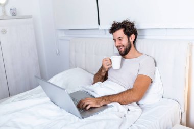 Kafkasyalı genç bir adamın sabah kahvesini içtiği, yatağında oturduğu, laptopuyla çalıştığı ve kahve içtiği, incelenmeye hazırlandığı, internette sörf yapmak için dizüstü bilgisayarını kullandığı görüntüsü..
