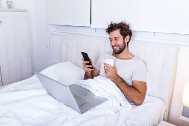 İşkolik konseptte cep telefonu kullanarak, sabahları evde çalışarak ve kahve içerek bilgisayarla mutlu ve rahat bir şekilde çalışan genç işadamı.