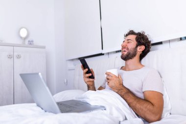 İşkolik konseptte cep telefonu kullanarak, sabahları evde çalışarak ve kahve içerek bilgisayarla mutlu ve rahat bir şekilde çalışan genç işadamı.