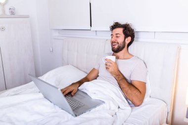 Kafkasyalı genç bir adamın sabah kahvesini içtiği, yatağında oturduğu, laptopuyla çalıştığı ve kahve içtiği, incelenmeye hazırlandığı, internette sörf yapmak için dizüstü bilgisayarını kullandığı görüntüsü..