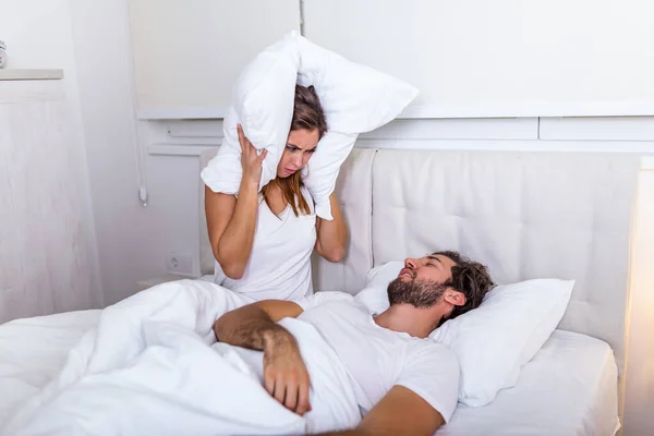 Young couple have problem with man's snoring. Heterosexual couple in bed, man sleeps and snoring with mouth open, while a tired woman irritated by snoring sitting on bed with a pillow on her head.