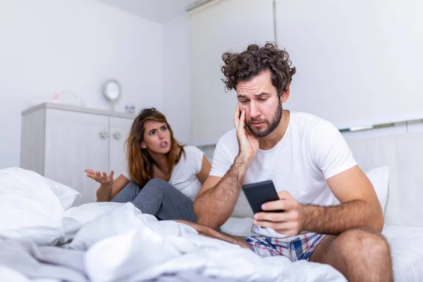 Young couple is sitting on the bed. The guy is looking at something on his smart phone. The girl is offended at him. She is looking at him annoyed and frustrated by boyfriend on the phone