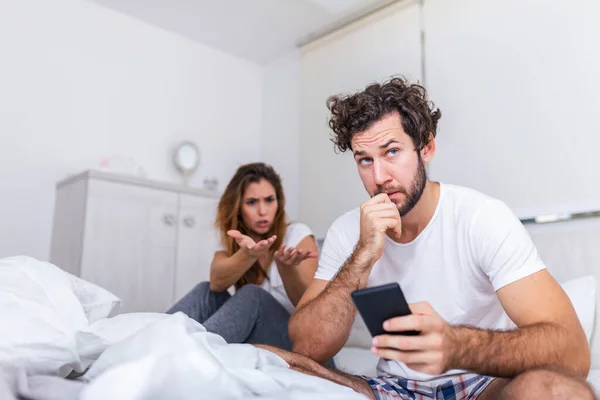 Young couple is sitting on the bed. The guy is looking at something on his smart phone. The girl is offended at him. She is looking at him annoyed and frustrated by boyfriend on the phone
