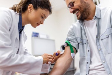 Kadın doktor üniformasının kan testi hazırlıkları beyaz parlak odadaki masada. Hemşire hastanın kolundaki damarı boş bir tüple deliyor..