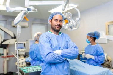 Hastanedeki ameliyathanede kolları çapraz duran erkek cerrahın portresi. Takım cerrahları ameliyat yapıyor, orta yaşlı doktor kameraya bakıyor, modern bir ameliyathanede.