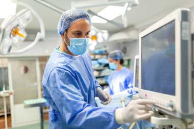Anesthetist Working In Operating Theatre Wearing Protecive Gear checking monitors while sedating patient before surgical procedure in hospital clipart