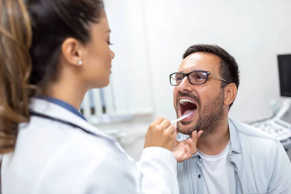 A young man sits on an exam table across from his doctor. The doctor reaches forward with a tongue depressor as the man looks up and sticks out his tongue.