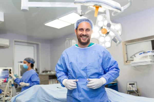 Portrait of male nurse surgeon OR staff member dressed in surgical scrubs gown mask and hair net in hospital operating room theater making eye contact smiling pleased happy looking at camera