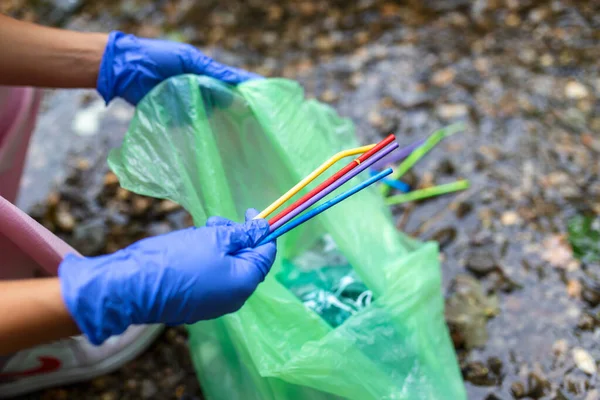 Hand of woman picking up plastic straws into garbage bags while cleaning small river. Volunteering, charity, people, ecology concept. Closeup volunteer collecting plastic trash in forest.