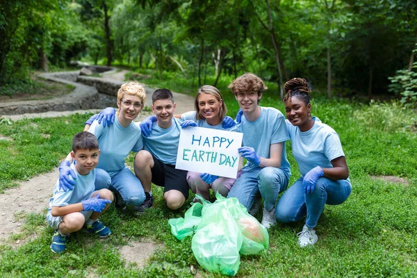 Diverse Group of People Picking Up Trash in The Park Volunteer Community Service. Happy international volunteers holding placard with \'happy Earth day\' message.