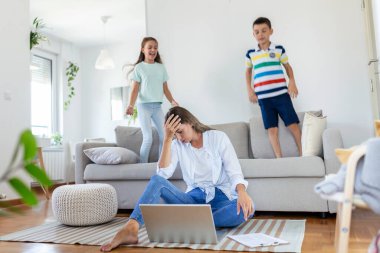 Küçük çocuklar kanepede zıplayıp eğlenirken ve gürültü yaparken genç anne yerde oturup dizüstü bilgisayarla ve belgelerle çalışıyor.