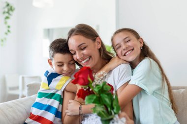 Elinde gül buketi olan genç bir anne gülüyor, oğluna sarılıyor ve elinde bir kartla korkunç bir kız, mutfakta tatilde annesini tebrik ediyor.