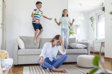 Küçük çocuklar kanepede zıplayıp eğlenirken ve gürültü yaparken genç anne yerde oturup dizüstü bilgisayarla ve belgelerle çalışıyor.