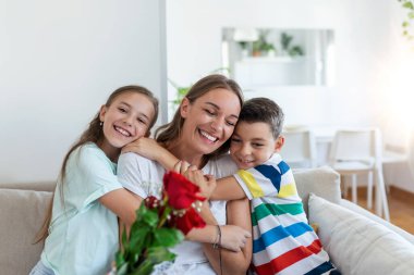 Elinde gül buketi olan genç bir anne gülüyor, oğluna sarılıyor ve elinde bir kartla korkunç bir kız, mutfakta tatilde annesini tebrik ediyor.
