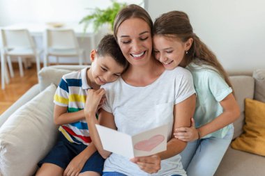 Mutlu küçük anne ve çocuklar kanepede oturup dileklerini okurken kucaklaşıyorlar ve evde sunulan kartpostalda tatille kutluyorlar.