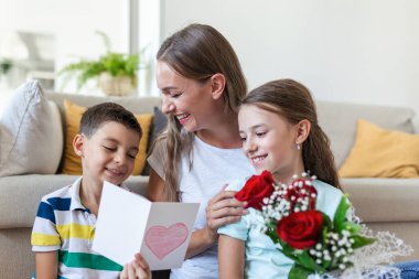 Elinde gül buketi olan genç bir anne gülüyor, oğluna sarılıyor ve elinde kart ve güllerle korkunç bir kız evde tatil kutlamalarında annesini tebrik ediyor. Anneler Günü