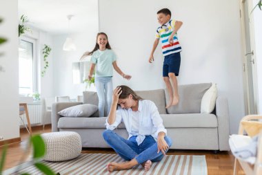 Küçük çocuklar kanepede zıplayıp eğlenirken ve gürültü yaparken genç anne yerde oturup dizüstü bilgisayarla ve belgelerle çalışıyor.
