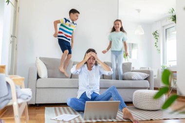 Küçük çocuklar kanepede zıplayıp eğlenirken ve gürültü yaparken genç anne yerde oturup dizüstü bilgisayarla ve belgelerle çalışıyor.