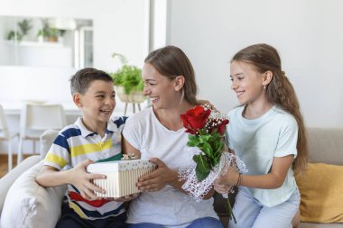 Mutlu çocuklar annelerine çiçek veriyorlar. Anneler Günün kutlu olsun! Çocuklar erkek ve kız gülümseyen anneyi kutlar, ona bir buket gül ve bayram kutlaması sırasında bir hediye kutusu verir.