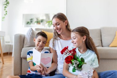 Elinde gül buketi olan genç bir anne gülüyor, oğluna sarılıyor ve elinde kart ve güllerle korkunç bir kız evde tatil kutlamalarında annesini tebrik ediyor. Anneler Günü