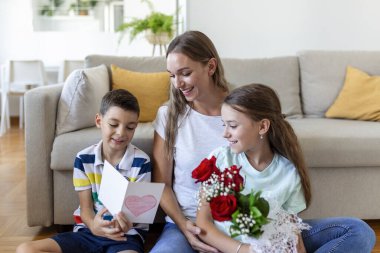 Elinde gül buketi olan genç bir anne gülüyor, oğluna sarılıyor ve elinde kart ve güllerle korkunç bir kız evde tatil kutlamalarında annesini tebrik ediyor. Anneler Günü