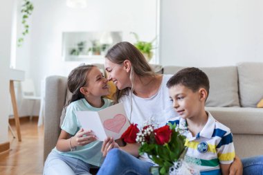 Elinde gül buketi olan genç bir anne gülüyor, oğluna sarılıyor ve elinde kart ve güllerle korkunç bir kız evde tatil kutlamalarında annesini tebrik ediyor. Anneler Günü