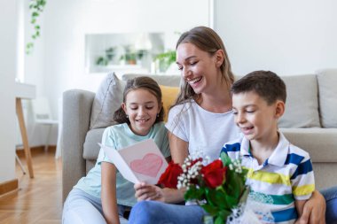Elinde gül buketi olan genç bir anne gülüyor, oğluna sarılıyor ve elinde kart ve güllerle korkunç bir kız evde tatil kutlamalarında annesini tebrik ediyor. Anneler Günü
